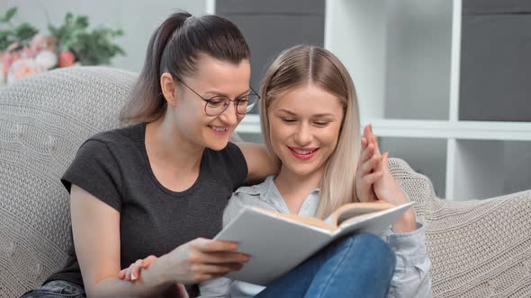 Two Smiling Homosexual Young Woman Reading Book Together on Couch Feeling Love and Tenderness