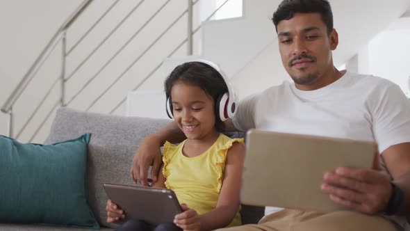 Happy hispanic father and daughter sitting on sofa using tablets