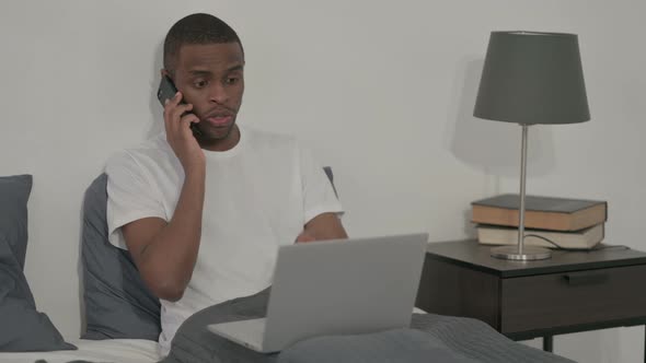 African Man with Laptop Talking on Smartphone Sitting in Bed