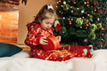 Little girl opens a Christmas present near the Christmas tree - PhotoDune Item for Sale