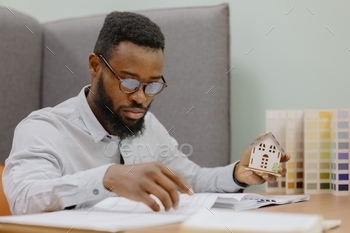 erior designer in his office. a black man creates a design project of a house or apartment for a customer. An African man is a young and handsome professional man holding a miniature model of a house in his hands. buying or selling a real estate apartment or house. sales and mortgage agent. a man with glasses a designer or designer makes drawings of a project of a residential house or commercial premises