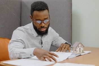 erior designer in his office. an African American man creates a design project of a house or apartment for a customer. An African man is a young and handsome professional man holding a miniature model of a house in his hands. buying or selling a real estate apartment or house. sales and mortgage agent. a man with glasses a designer or designer makes drawings of a project of a residential house or commercial premises