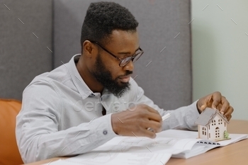 erior designer in his office. an African American man creates a design project of a house or apartment for a customer. An African man is a young and handsome professional man holding a miniature model of a house in his hands. buying or selling a real estate apartment or house. sales and mortgage agent. a man with glasses a designer or designer makes drawings of a project of a residential house or commercial premises