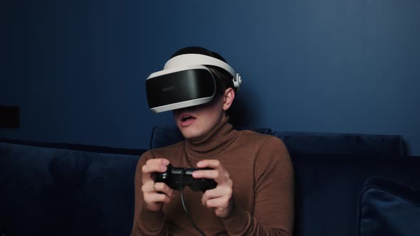 Attractive Caucasian Man Hold Joystick and Wear Virtual Reality Glasses Headset Helmet on Sofa at