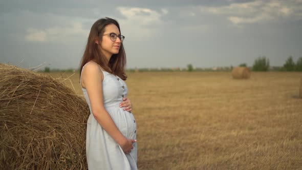 A Young Pregnant Woman Stands Near a Haystack in a Field