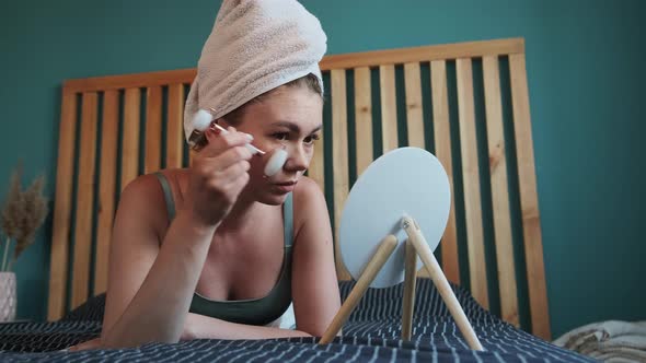 Young Woman Using Jade Facial Roller for Face Massage Lying on Bed in Bedroom Looking in the Mirror