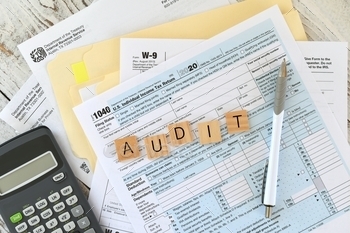  an IRS envelope alongside a calculator with the word AUDIT spelled out in wooden letter tiles. Having your income tax filings audited to make sure you filed correctly and claimed everything you were supposed to.
