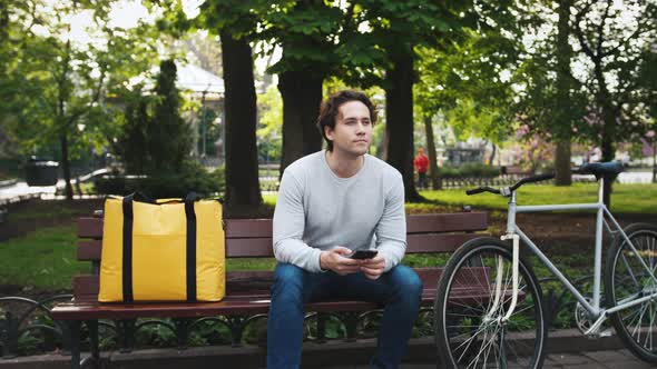 Portrait of Caucasian Delivery Man Sitting on Bench in Central Park and Using Smartphone with Yellow