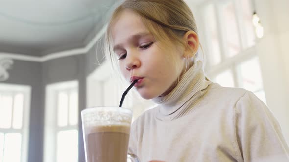 a Little Blonde Girl Drinks Cocoa From a Glass Through a Straw