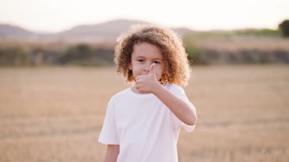 Cute little boy with dreadlocks giving positive thumps up sign on agricultural field during sunset