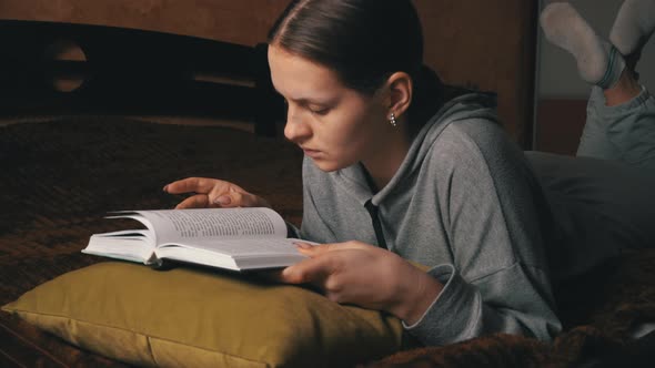 Cute Girl Reads and Leafs Through a Book While Lying Comfortably in Bed