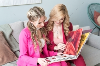  choose a hair color for dyeing. Two young women women leaf through an album with samples of hair dyes. Millennials girls are sitting on the couch watching a large magazine together. Caucasian girls are resting on the couch in the living room. Women of European appearance with long blond hair chat while sitting on the couch in the room. Kaaral. Baco.