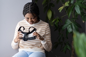 VR glasses in hands while relaxing near window at home, young african american lady testing modern virtual reality headset for video games