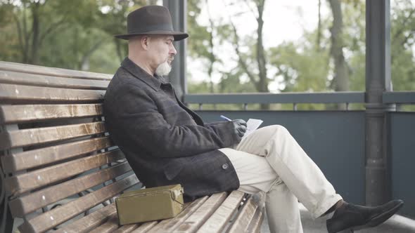 Side View of Elegant Undercover Agent or Spy Sitting on Bench in Cloudy Spring or Autumn Park and
