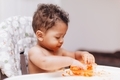 Diverse baby at home eating jar carrots in high chair while making a mess - PhotoDune Item for Sale