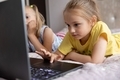 Two sisters 7-8 years old playing a game on a laptop lying on the bed in the room. - PhotoDune Item for Sale