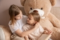 Two sisters are sitting in a room on a bed with a big teddy bear and reading a fairy tale in a book. - PhotoDune Item for Sale