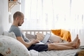 A young father working from home on laptop sitting on the bed during daytime sleeping of the baby. - PhotoDune Item for Sale