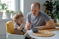 Close up of a father and child during lunch. A young dad supporting his baby toddler in self-feeding - PhotoDune Item for Sale