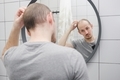 Stressed man with hair loss problem critically looking at mirror in the bathroom holding hairclipper - PhotoDune Item for Sale
