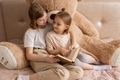 Two sisters are sitting in a room on a bed with a big teddy bear and reading a fairy tale in a book. - PhotoDune Item for Sale