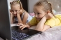 Two sisters 7-8 years old playing a game on a laptop lying on the bed in the room. - PhotoDune Item for Sale