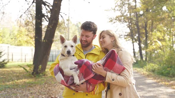 Young Married Couple Holds Their Pet Dog Wrapped in a Checkered Blanket Outside in a Park