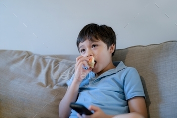  on TV, Mixed race school kid relaxing at home after back from school, Positive child sitting on sofa eating fruit for snack in living room.