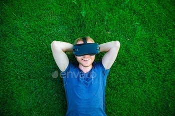 man enjoying virtual reality glasses headset or 3d spectacles lying on the green lawn in city park