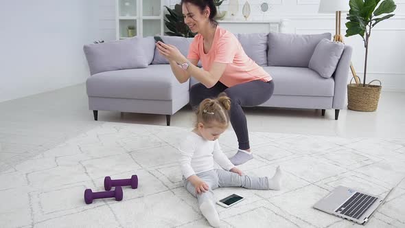 Girl Sitting on the Floor and Watching Cartoon on Phone while Her Mother Doing Squat Exercises