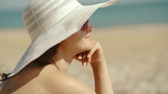 A Young Girl in a White Hat Sits on the Seashore and Looks Into the Distance