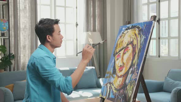 Side View Of An Asian Artist Man Thinking Before Painting A Girl's Hair On The Canvas