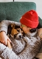 Young woman in an orange beanie and comfy sweater snuggling and kissing a comfy puppy dog on a couch - PhotoDune Item for Sale