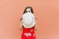 A woman in a summer sun dress standing against a simple orange wall covering her face with a hat.  - PhotoDune Item for Sale