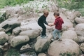 A young boy helping another little boy across some big rocks outside in nature.  - PhotoDune Item for Sale