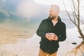 A man with a beard in a cozy sweater with a cup of coffee camping at the lake.  - PhotoDune Item for Sale