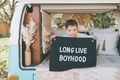 A happy little boy in the back of a camper van holding up a sign that says 'long live boyhood'.  - PhotoDune Item for Sale