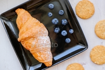 Croissant on a black square plate with blueberries and whole grain cookies. Flat lay. Cozy breakfast