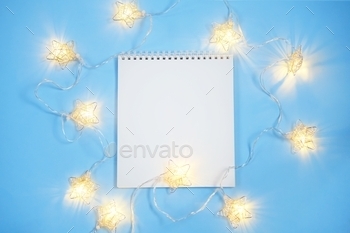 Blank page of a notepad and Christmas decorative lights on light blue background