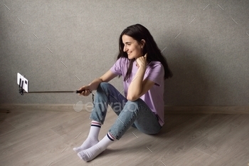 A young girl is sitting on the floor and holding a selfie stick with her phone to take a photo
