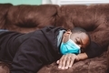 African American man at home in blue mask laying on coach looking sick. - PhotoDune Item for Sale