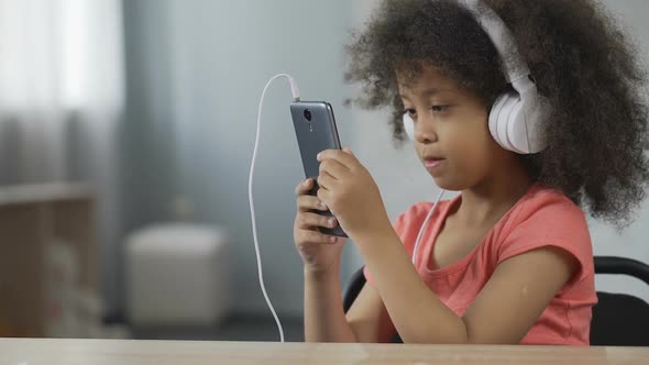 Pretty Afro-American Child Wearing Earphones and Listening to Music on Cellphone