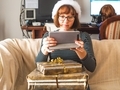 Young woman with stack of gifts looking at her tablet sitting on couch at home in living room - PhotoDune Item for Sale