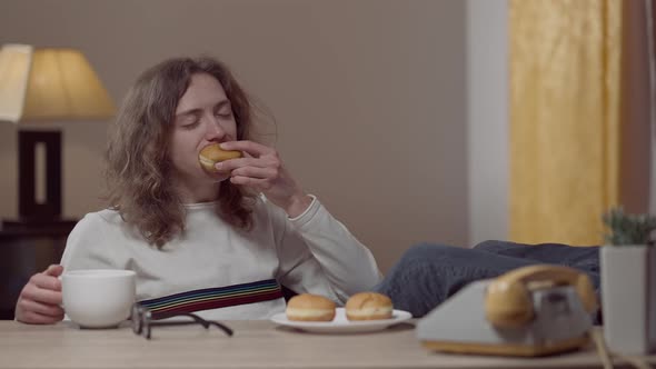 Carefree Retro Man Eating Donuts and Drinking Tea or Coffee Sitting with Feet on Table Indoors