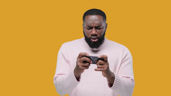Black Young Man Enthusiastically Playing Game on Smartphone on Yellow Background