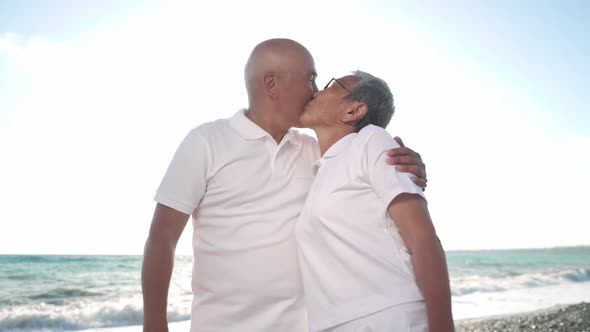 Excited Senior Asian Woman and Adult Man Kissing in Sunlight Standing on Mediterranean Sea Beach at