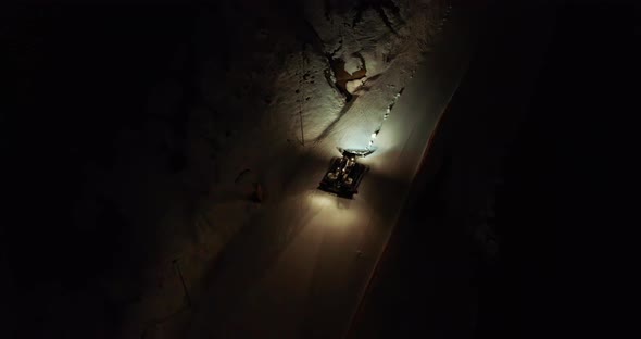 Orange Illuminated Snowcat Drives Up Along White Snowy Hill on Track in Evening Darkness