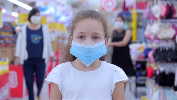 Cute Little Girl From an Epidemic of Coronaviruses or Viruses Looks at the Camera Amid Masked People