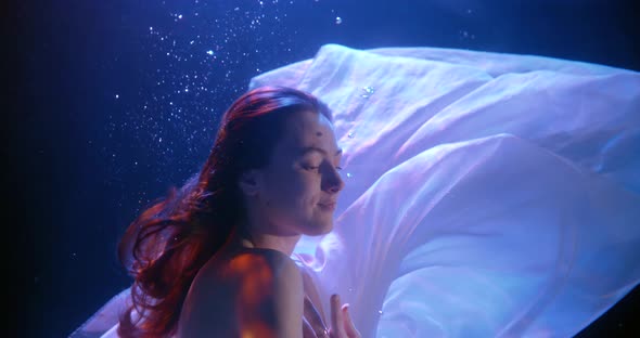 Young Woman Is Dancing Underwater with Open Eyes, White Chiffon Fabric Is Floating Around