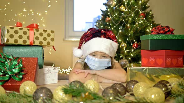 Sad Girl 6-7 Years Old Wearing a Santa Hat and a Protective Mask Sits Alone